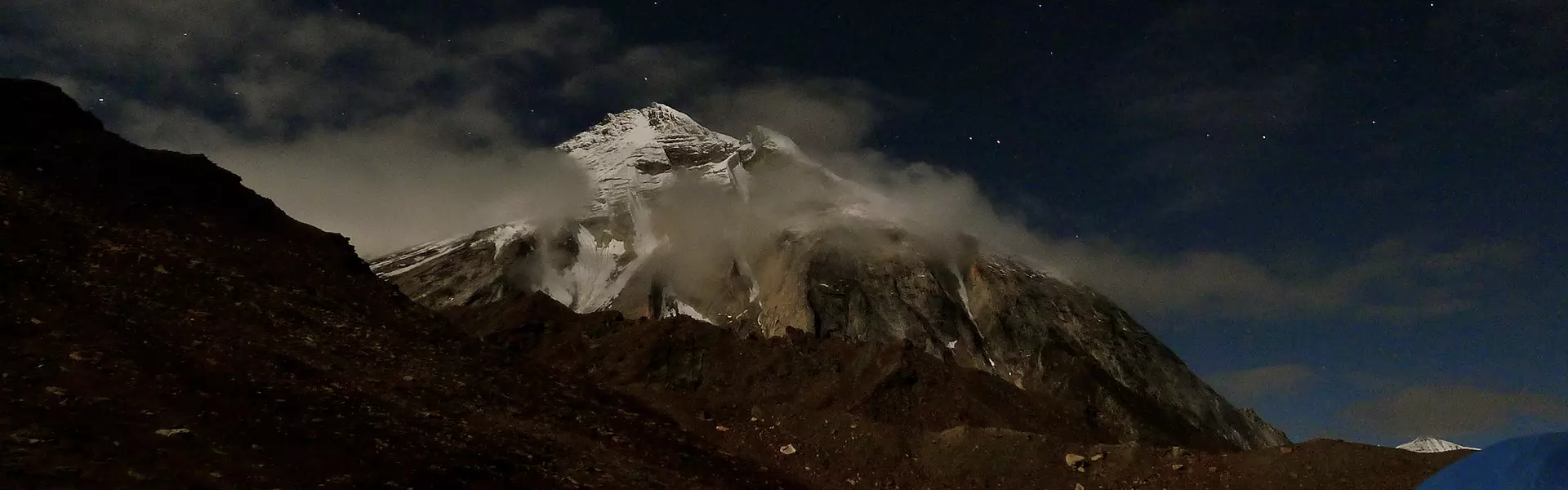 Twee glimlachende bergbeklimmers in blauwe jassen en met zonnebrillen staan dicht bij elkaar op een besneeuwde berg; het felle zonlicht weerkaatst op hun brillen, met een helderblauwe lucht achter hen